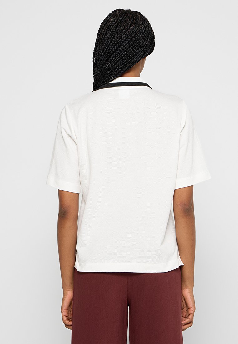 Haut blanc à manches courtes en tissu texturé, avec un col noir et des fentes sur les côtés au niveau de l'ourlet, porté avec un pantalon bordeaux.