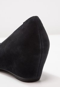 Chaussure à talon compensé en daim noir avec un bout pointu et une finition texturée, présentant une surface extérieure lisse et un léger éclat.