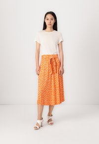 Oranger Midi-Rock mit weißem Blattmuster, leichtes Material, elastischer Bund mit Bindedetail, kombiniert mit einem schlichten weißen T-Shirt und weißen Absatzschuhen.