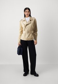 Giacca in pelle beige aderente con cintura, chiusura a zip e spalline, abbinata a pantaloni neri larghi e una pochette a trama blu navy.