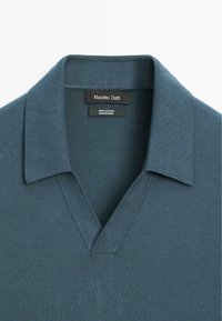 Bawełnowy sweter w ciemnym odcieniu zieleni z dekoltem w kształcie V i złożonym kołnierzem. Posiada gładką teksturę oraz subtelne prążkowane wykończenia. Widoczna etykieta marki.