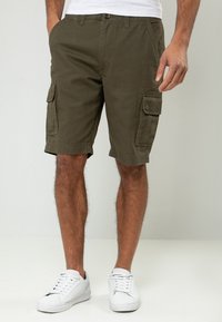 Shorts cargo vert olive en tissu résistant, avec plusieurs poches et des détails cousus. Portés avec des baskets blanches.