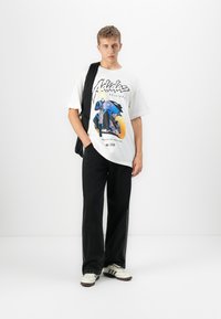 Ung mann kledd i en grafisk hvit t-skjorte, svarte vide bukser, hvite sneakers, og bærende en svart jakke over den ene skulderen.