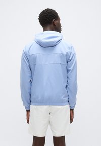 Casaco corta-vento azul claro com capuz, punhos e bainha elásticos. Tecido liso e resistente à água. Costuras horizontais em contraste nas costas.