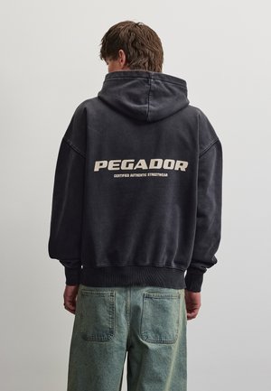 Homme portant un sweat à capuche noir avec le texte « PEGADOR Certified Authentic Streetwear » dans le dos et un jean vert délavé.