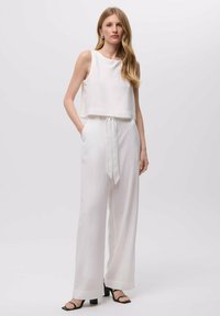 Bílý bezrukávový cropped top doplněný volnými bílými kalhotami s páskem na zavázání v pase. Modelka má na sobě černé sandály na podpatku.