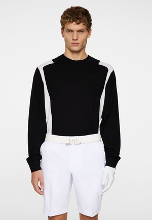 Sorte langærmede skjorte med hvide mesh-detaljer, båret med hvide shorts og et hvidt bælte. Modellen holder en hvid handske i højre hånd.