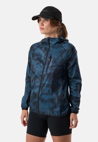 Femme portant une casquette noire, une veste à capuche zippée bleu camouflage et un short noir, debout et regardant sur le côté sur un fond blanc.