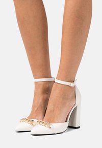 Chaussures à talons hauts blanches avec un bout pointu, une bride à la cheville et un détail de chaîne dorée à l'avant. Texture en cuir lisse et talon épais.