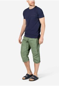 Marineblaue Baumwoll-T-Shirt mit einer Brusttasche, kombiniert mit knielangen grünen Cargo-Shorts mit Taschen und Reißverschlussakzenten. Schwarze Sandalen.