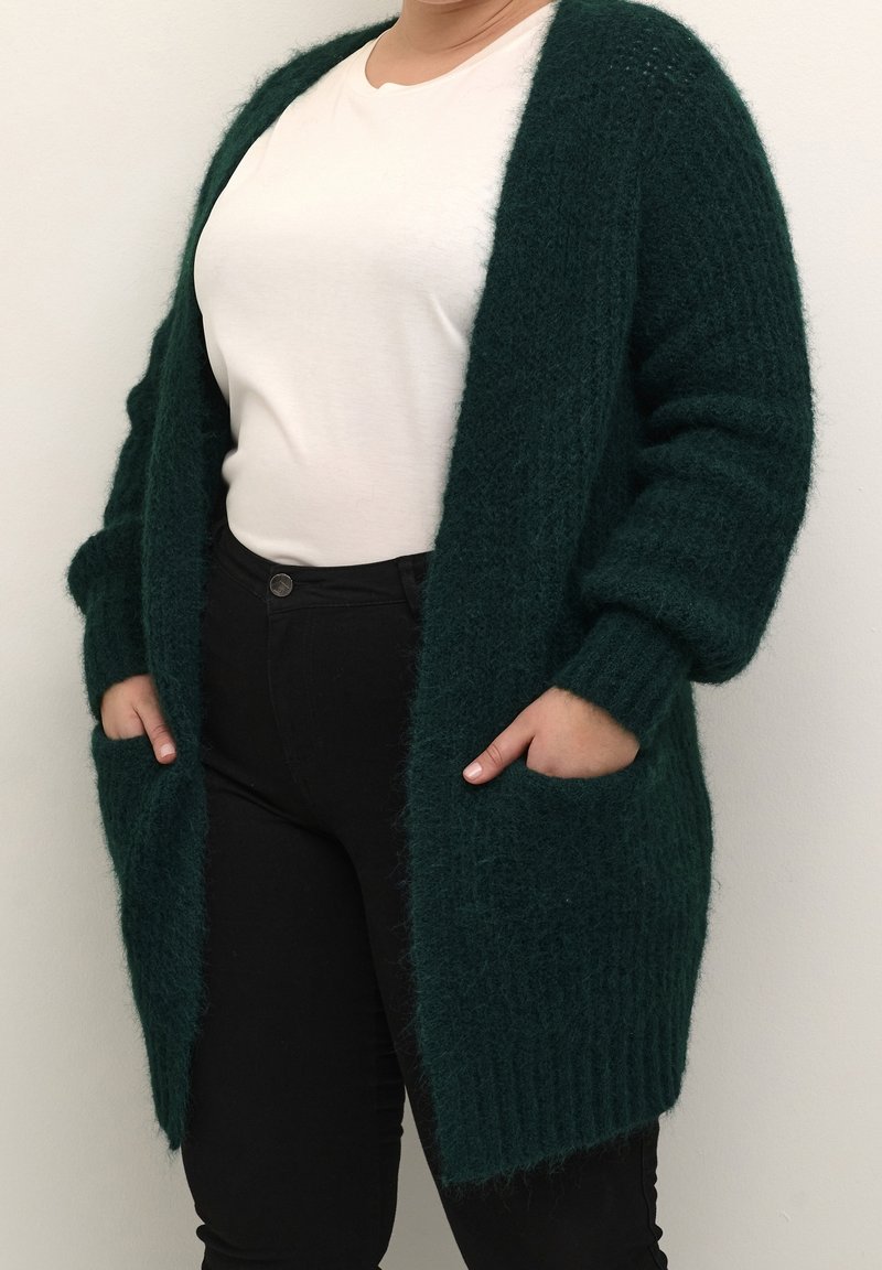 Lang, åben-front cardigan lavet af fuzzy, mørkegrøn materiale. Har ribbede manchetter og lommer; parret med en lysfarvet skjorte og sorte bukser.