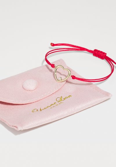 Sarkans pītā aukla aproce ar zelta sirds piekariņu un mazu dimanta akcentu, pasniegta rozā zamša maisiņā ar zelta zīmolu.