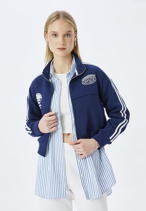 Marineblauwe zip-up jack met witte strepen en logo-accenten, gedragen over een blauw-wit gestreept shirt. Witte tailleband, neutrale achtergrond.
