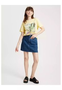 Żółta koszulka graficzna z wzorem palmy, w zestawie z niebieską dżinsową spódnicą. Czarne sandały na platformie dopełniają stylizację.