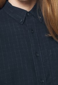 Tmavě modrá košile s knoflíky a jemným kostkovaným vzorem, s překládaným límcem, texturovanou tkaninou a černými knoflíky na přední straně.