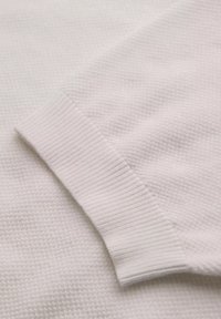 Bílá pletenina s texturovaným povrchem a žebrovaným manžetem. Celkový design je jednoduchý a čistý, zdůrazňující měkkost materiálu.