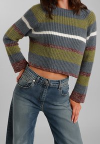 Maglione grigio corto con texture a coste, caratterizzato da righe orizzontali in verde oliva, bianco e bordeaux. Indossato con jeans azzurri chiari.