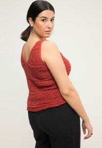 Krajkový tank top v rezavé barvě s výstřihem do V a složitými vlnitými vzory, doplněný černými kalhotami, které ukazují texturovaný design.