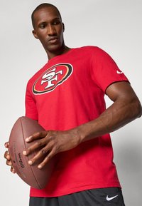 Red cotton t-shirt with a bold black and white San Francisco logo, paired with a brown football, featuring a textured surface and grip.