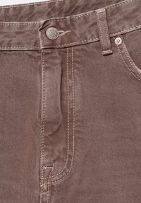 Gros plan d'un jean en denim marron montrant la ceinture, la boucle de ceinture, le bouton métallique, le rivet, la poche avant et les coutures détaillées.