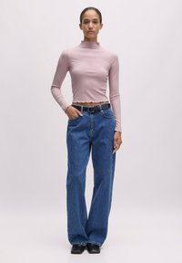 Langärmliges mauvefarbendes Rollkragenoberteil mit gewelltem Saum, kombiniert mit weit geschnittenen blauen Denim-Jeans und einem schwarzen Gürtel mit einer silbernen Schnalle.