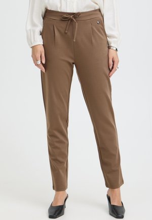 Femme portant un pantalon marron fuselé avec une taille à cordon, associé à des mules à talons noires et un haut blanc à manches longues.