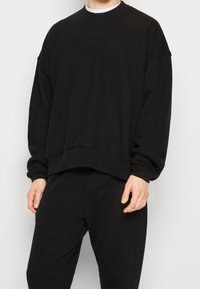 Černá oversize mikina s kulatým výstřihem, texturovanou tkaninou, spadlými rameny a žebrovaným lemem. Nosená s odpovídajícími černými kalhotami.