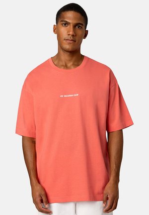 Korallefarbene Rundhalsausschnitt-T-Shirt aus weichem Baumwollstoff mit kurzen Ärmeln, mit weißem Text "ON VACATION CLUB" quer über der Brust.