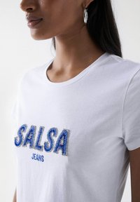 Biała bawełniana koszulka z okrągłym dekoltem, krótkimi rękawami i lekko elastycznym materiałem, z napisem "SALSA" w wypukłym, niebieskim i srebrnym haftem.