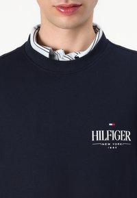 Marineblauwe sweatshirt met geribbelde ronde hals, met het logo "HILFIGER NEW YORK 1985" in het wit. Gelaagd met een lichte gestreepte kraag eronder.