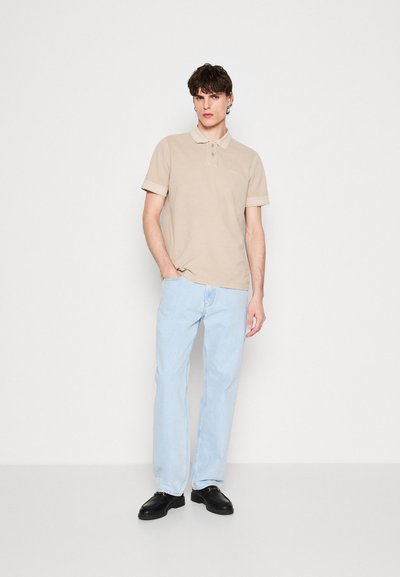 Polo beige à manches courtes, avec trois boutons et un détail de logo ; associé à un jean droit bleu clair et des chaussures noires.