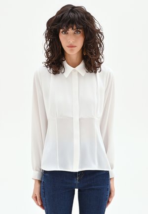 Femme aux cheveux bouclés foncés portant des boucles d'oreilles en or, un chemisier blanc à manches longues boutonné et un jean bleu foncé sur un fond blanc uni.