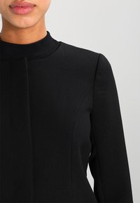 Mujer vestida con una chaqueta negra ajustada, con un cuello alto y detalles de costuras verticales, mostrada de la mitad de la cara a la mitad del torso contra un fondo gris.