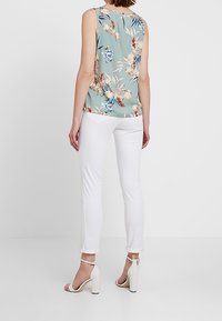 Blusa sin mangas con estampado floral en verde claro, pantalones ajustados blancos y sandalias de tacón blancas con tiras en el tobillo. Vista posterior.