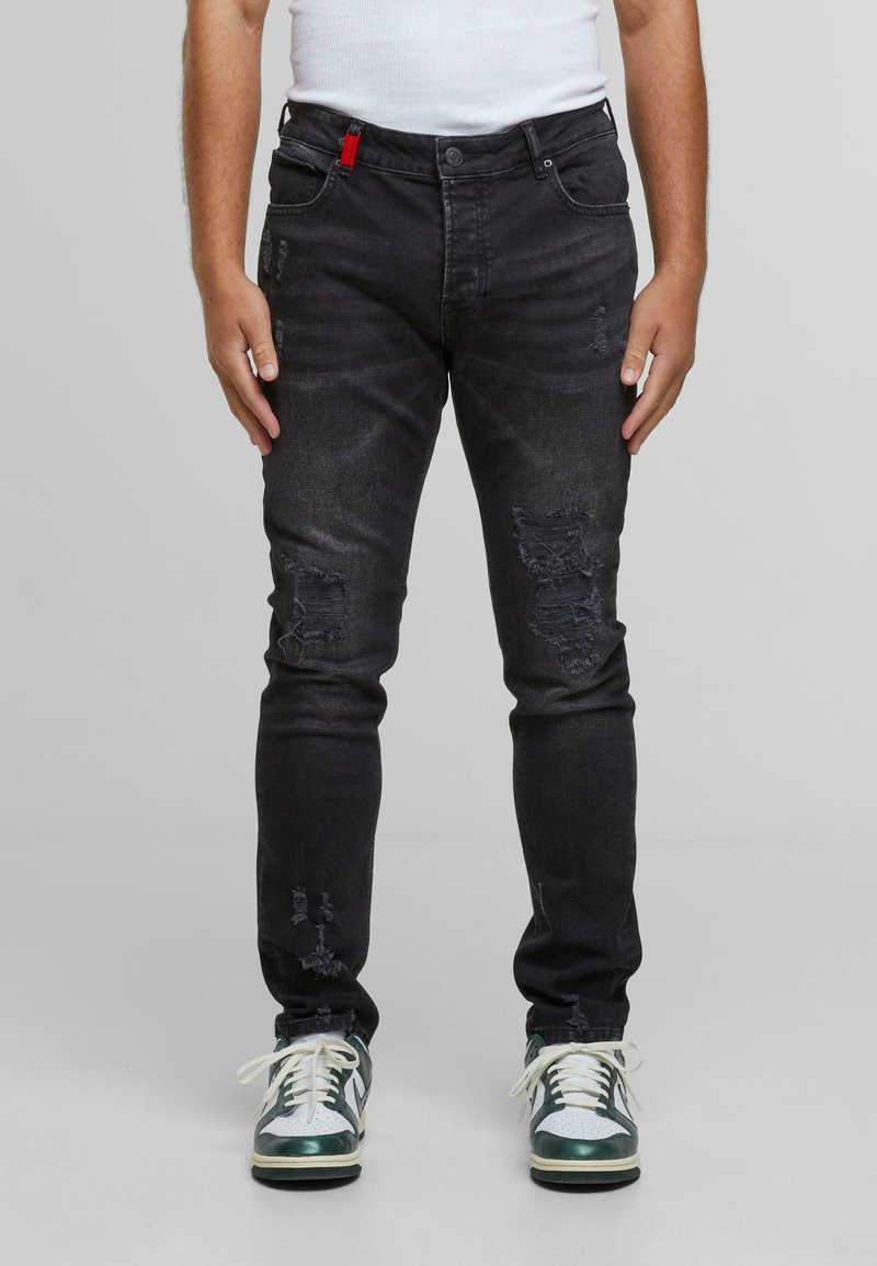 Man in zwarte versleten slim-fit jeans en wit-groene sneakers, staande tegen een effen lichtgrijze achtergrond.