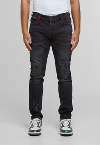 Vyras vilkintis juodais nudėvėtais siaurais džinsais ir baltais-žaliais sportbačiais, stovintis prieš paprastą šviesiai pilką foną.