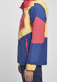 Kolorowa kurtka wiatrówka z żółtymi, niebieskimi i czerwonymi panelami. Posiada zamek błyskawiczny z przodu, regulowany kaptur oraz elastyczne mankiety dla lepszego dopasowania.