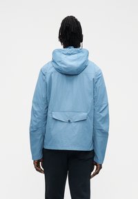 Casaco azul claro com capuz e corte descontraído, apresentando um bolso nas costas com fecho de botão e tecido texturizado. O design das mangas inclui espaço amplo.