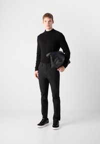 Suéter de punto negro, pantalones negros ajustados y zapatillas negras con suelas blancas. El modelo sostiene un bolso de cuero negro. Diseño simple, acentos mínimos.