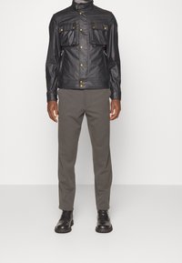 Svart läderjacka med hög krage, två bröstfickor och gyllene detaljer, tillsammans med grå byxor och svarta snörkängor.