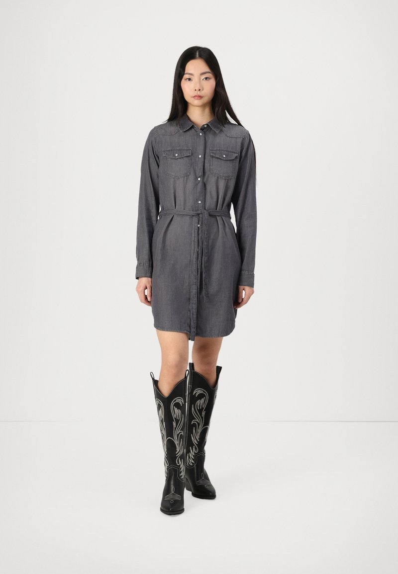Robe chemise en denim gris avec une taille ceinturée, des manches longues et deux poches poitrine, associée à des bottes de cowboy brodées noires surdimensionnées.