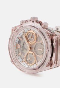 Et transparent pink har vækkeur med rund urskive, guldfarvede detaljer og tre undermetere. Har sølvfarvede timemarkører og logo.