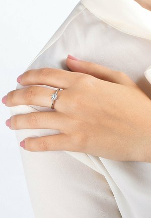 Silberring mit einem zentralen runden, klaren Edelstein, auf einem dünnen Band montiert. Hand ruht auf einem hellen Stoffärmel. Fingernägel in zartem Rosa lackiert.