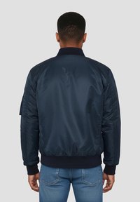 Homme portant une veste bomber bleu marine foncé et un jean bleu, vu de dos sur un fond gris clair uni.