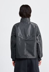 Mörkgrå, glänsande jacka med hög krage, oversized passform och dragsko vid ärmsluten. Har en utmärkande ryggdesign med vinklade sömmar.