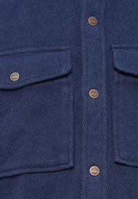 Tamno plavi flanel košulja s dva džepa na dojkama, brončanim gumbima s logotipom drveta i teksturiranim ribljim kostima.