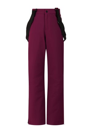 Jeune femme portant un haut à manches longues rose vif, un pantalon de neige violet avec bretelles noires, des lunettes de soleil roses et des bottes noires, debout sur un fond uni.