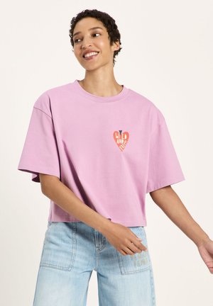 Junge Person lächelt, trägt ein locker sitzendes, hellviolettes bauchfreies T-Shirt mit einer kleinen roten Herz-Grafik und hellblaue hoch taillierte Jeans.