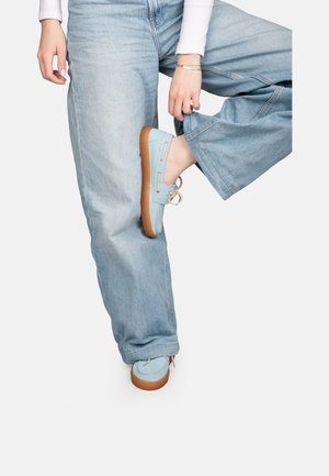 Persona con vaqueros de mezclilla azul claro y zapatos náuticos azul claro con suelas marrones, levantando una pierna doblada en la rodilla.