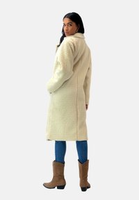 Abrigo de felpa texturizada beige con una silueta larga, bolsillos laterales y un cuello con muesca, combinado con vaqueros azules y botas de tobillo marrones.